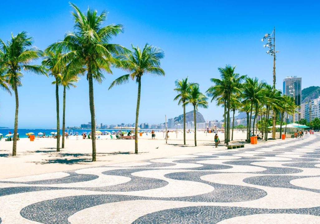 Sunny beach scene with palm trees lining a mosaic-patterned walkway. Blue skies, vibrant umbrellas, and distant mountains create a lively, tropical vibe.