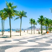 Sunny beach scene with palm trees lining a mosaic-patterned walkway. Blue skies, vibrant umbrellas, and distant mountains create a lively, tropical vibe.