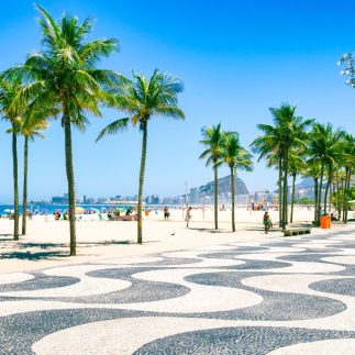 Sunny beach scene with palm trees lining a mosaic-patterned walkway. Blue skies, vibrant umbrellas, and distant mountains create a lively, tropical vibe.