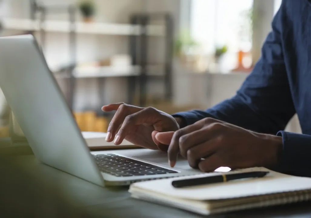 A person is typing on a laptop at a desk, with a notebook and pen nearby. The background shows shelves and a window.