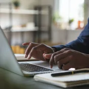 A person is typing on a laptop at a desk, with a notebook and pen nearby. The background shows shelves and a window.