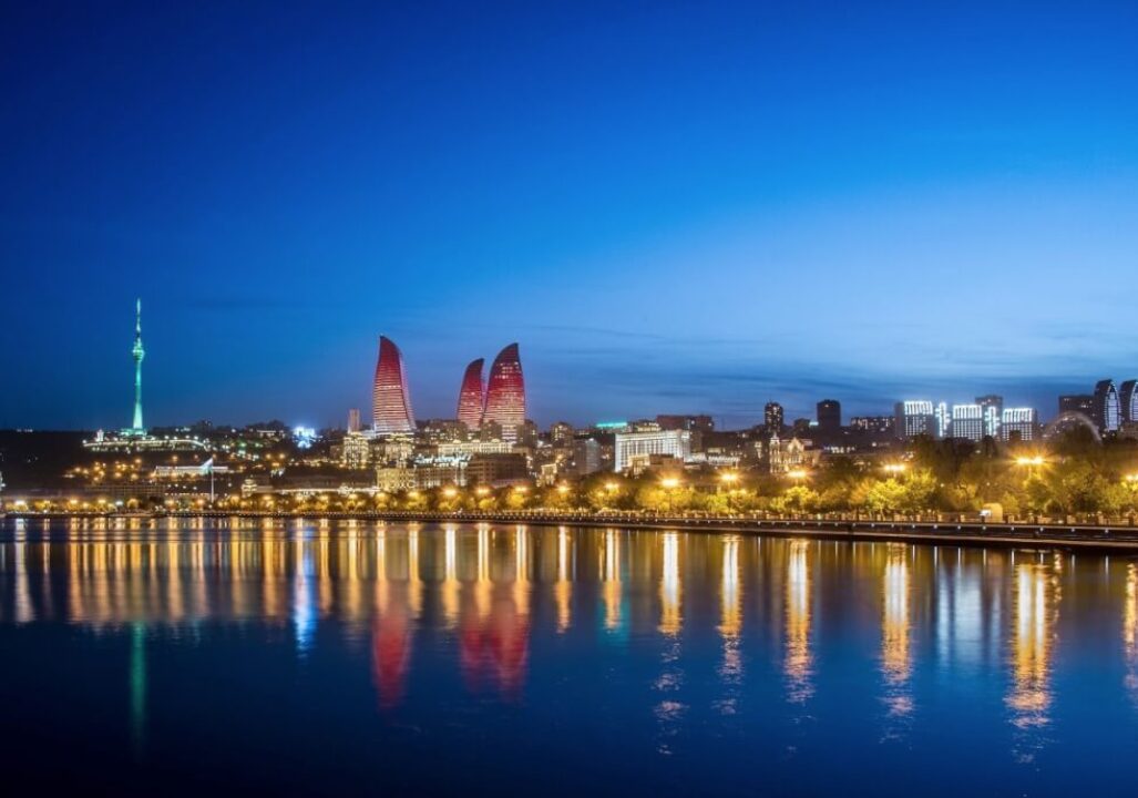 Nighttime cityscape of Baku with glowing Flame Towers, lit waterfront, and reflections on the water. A serene blue sky adds tranquility.