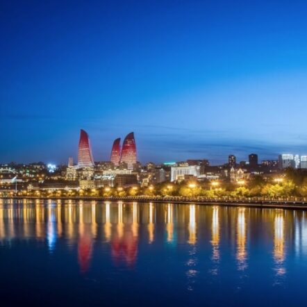 Nighttime cityscape of Baku with glowing Flame Towers, lit waterfront, and reflections on the water. A serene blue sky adds tranquility.