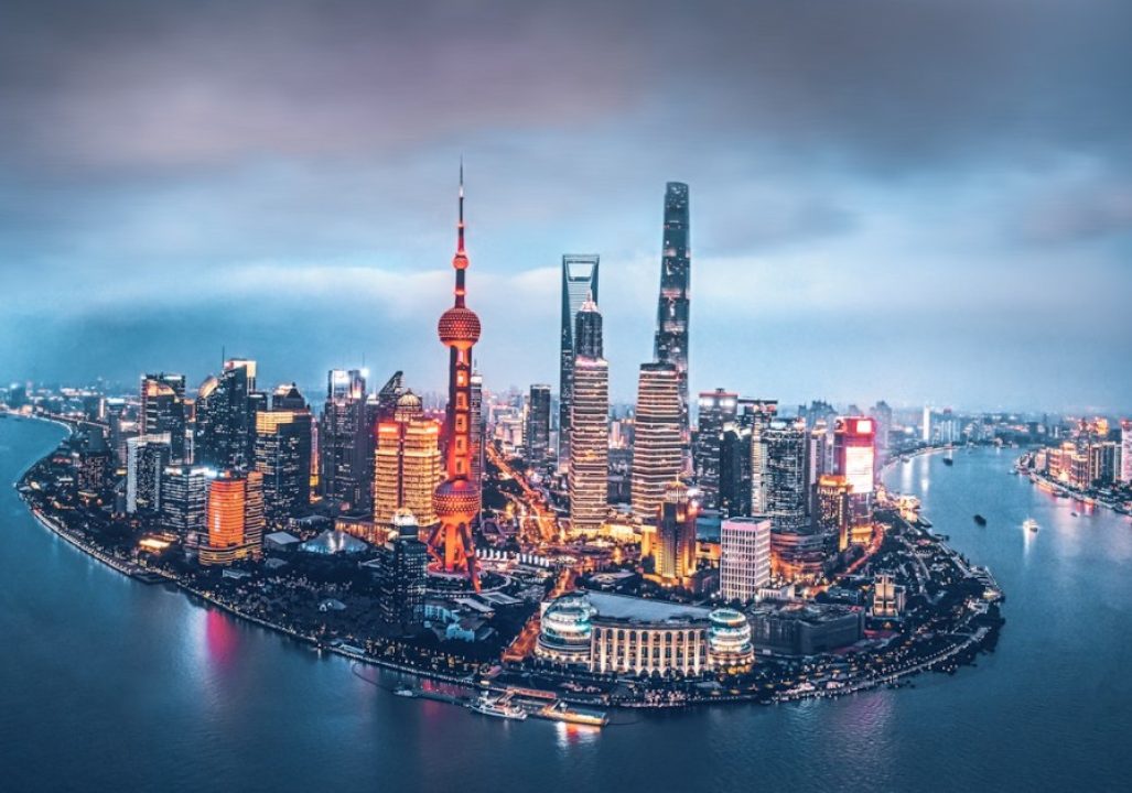 Aerial view of a vibrant city skyline at dusk, with illuminated skyscrapers, including a tower with a spherical design, beside a wide river under a cloudy sky.