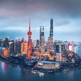 Aerial view of a vibrant city skyline at dusk, with illuminated skyscrapers, including a tower with a spherical design, beside a wide river under a cloudy sky.