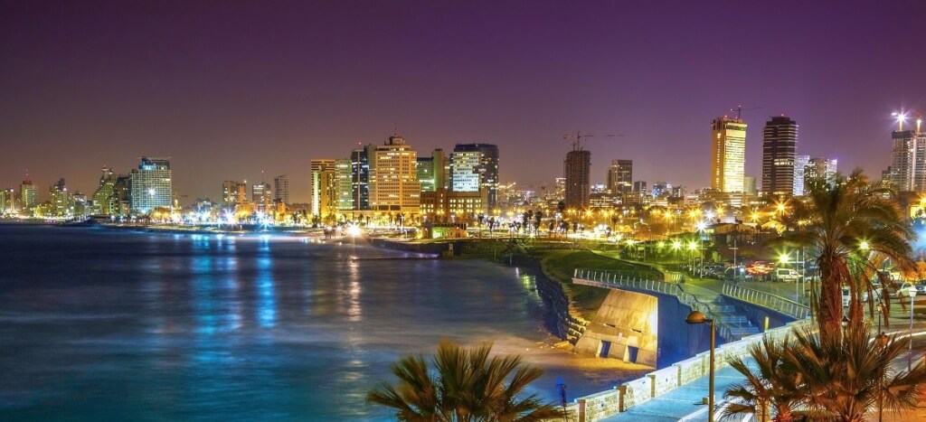 Illuminated modern skyline of Tel Aviv, Israel along its Mediterranean coastline at night, with palm trees in the foreground.