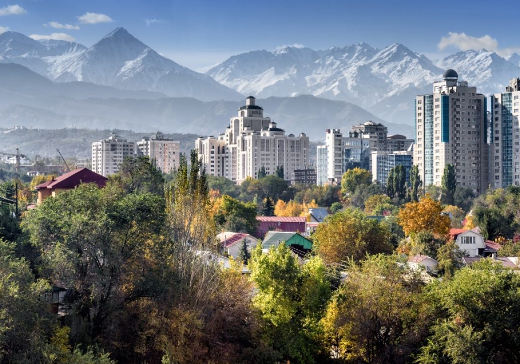 Cityscape with modern high-rise buildings framed by snow-capped mountains in the background. Foreground features lush greenery and colorful rooftops.