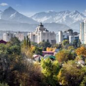 Cityscape with modern high-rise buildings framed by snow-capped mountains in the background. Foreground features lush greenery and colorful rooftops.