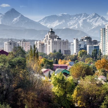 Cityscape with modern high-rise buildings framed by snow-capped mountains in the background. Foreground features lush greenery and colorful rooftops.