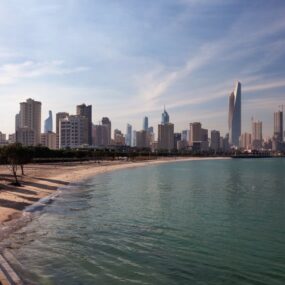 A Kuwait modern skyline along the coast, with a distinctive curved skyscraper and other high-rise buildings overlooking the Arabian Gulf.