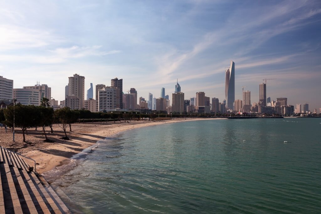 A Kuwait modern skyline along the coast, with a distinctive curved skyscraper and other high-rise buildings overlooking the Arabian Gulf.