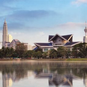 Kuala Lumpur, Malaysia featuring the Petronas Twin Towers and the Kuala Lumpur Tower in the background, with a foreground of modern architecture of the National Mosque or similar government buildings in front of a lake.