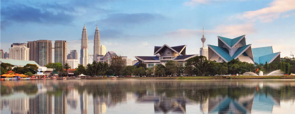 Kuala Lumpur, Malaysia featuring the Petronas Twin Towers and the Kuala Lumpur Tower in the background, with a foreground of modern architecture of the National Mosque or similar government buildings in front of a lake.