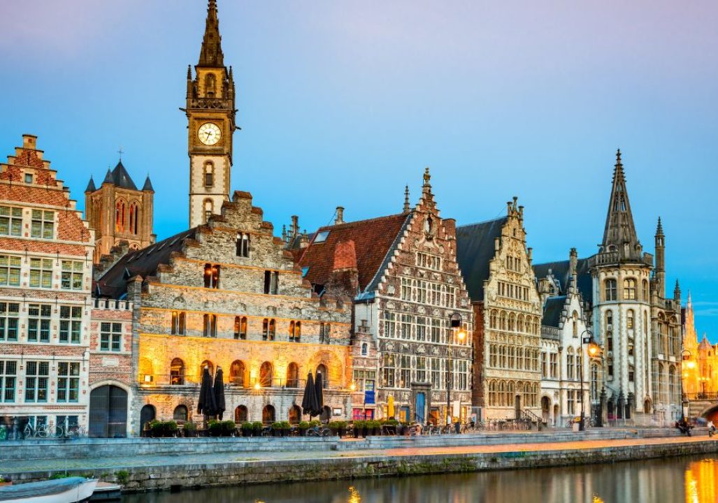 Historic European buildings line a canal at twilight, with warm lights illuminating their ornate facades. A tall clock tower rises prominently.