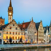 Historic European buildings line a canal at twilight, with warm lights illuminating their ornate facades. A tall clock tower rises prominently.