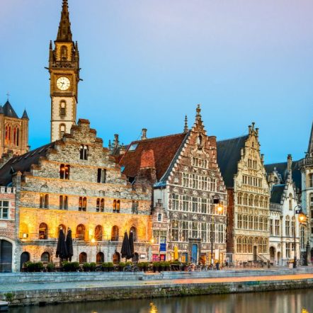 Historic European buildings line a canal at twilight, with warm lights illuminating their ornate facades. A tall clock tower rises prominently.