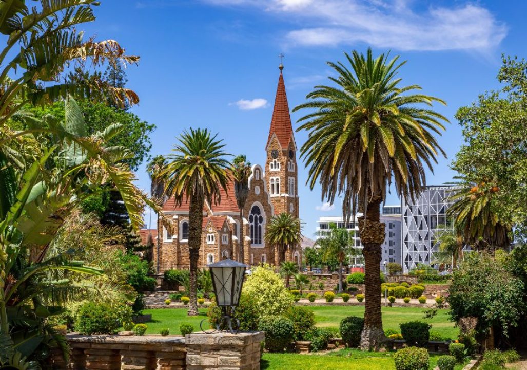 A picturesque church with a tall spire is framed by lush palm trees and vibrant greenery under a clear blue sky, conveying a serene and scenic atmosphere.