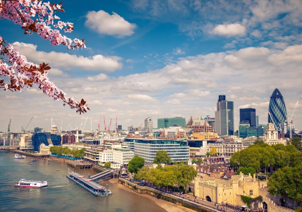 A view of London's skyline under a blue sky with clouds. Cherry blossoms frame the scene, and boats are on the Thames River. The Gherkin and city buildings are visible.