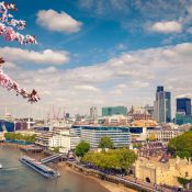 A view of London's skyline under a blue sky with clouds. Cherry blossoms frame the scene, and boats are on the Thames River. The Gherkin and city buildings are visible.