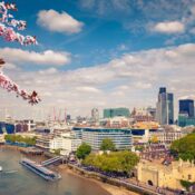 A view of London's skyline under a blue sky with clouds. Cherry blossoms frame the scene, and boats are on the Thames River. The Gherkin and city buildings are visible.