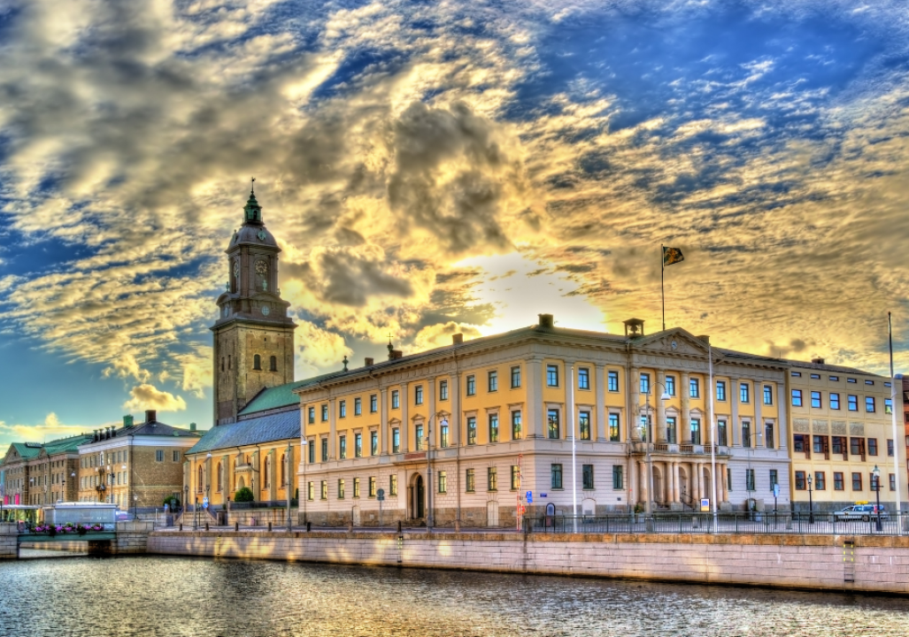 A historic building and church tower stand by a canal under a dramatic, cloudy sky at sunset in an urban cityscape.