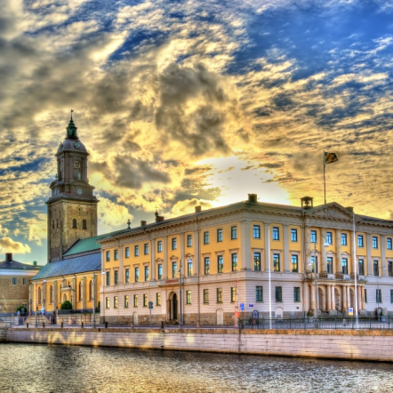 A historic building and church tower stand by a canal under a dramatic, cloudy sky at sunset in an urban cityscape.