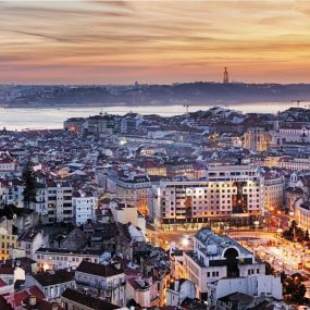 A panoramic view of a Portugal with many buildings, a castle on a hill to the left, and a large body of water with a bridge in the distance, all under a sunset sky.