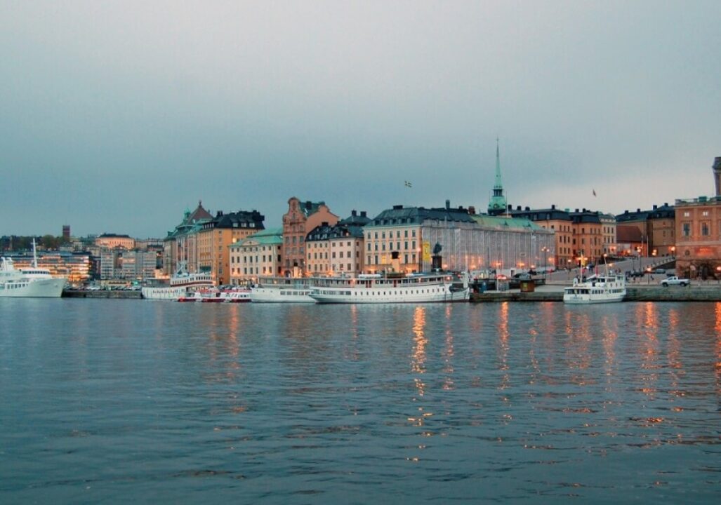 Sweden, features colorful historic buildings lining a waterfront with boats docked, under an overcast sky.