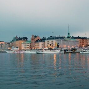 Sweden, features colorful historic buildings lining a waterfront with boats docked, under an overcast sky.