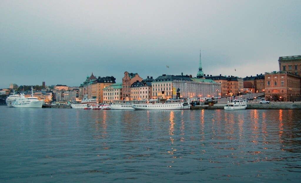 Sweden, features colorful historic buildings lining a waterfront with boats docked, under an overcast sky.