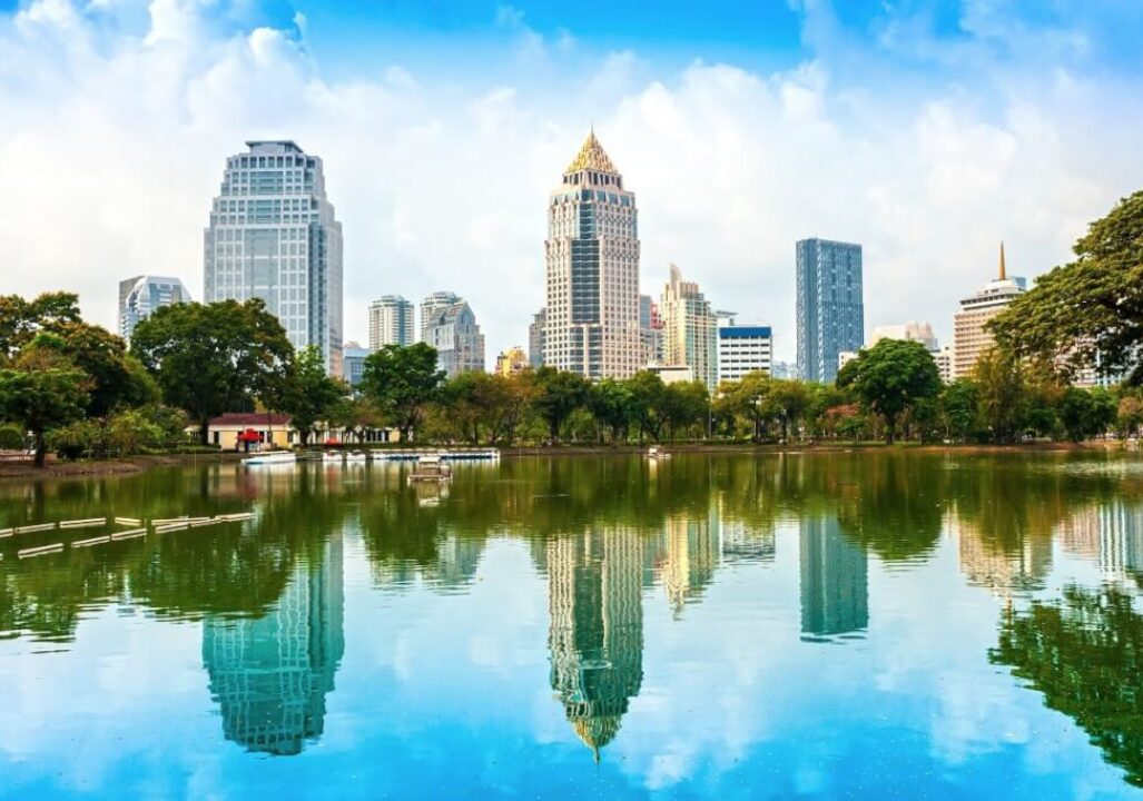 Thailand, shows a modern cityscape with numerous high-rise buildings reflected in a body of water, with green trees in the foreground.