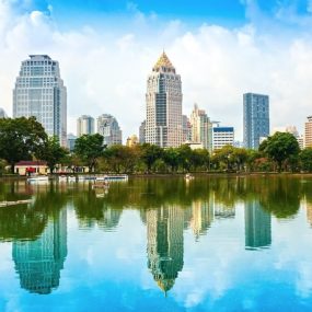 Thailand, shows a modern cityscape with numerous high-rise buildings reflected in a body of water, with green trees in the foreground.