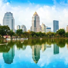 Thailand, shows a modern cityscape with numerous high-rise buildings reflected in a body of water, with green trees in the foreground.