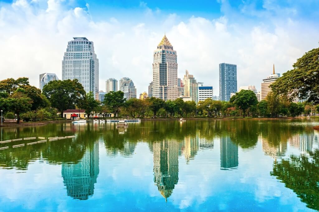 Thailand, shows a modern cityscape with numerous high-rise buildings reflected in a body of water, with green trees in the foreground.