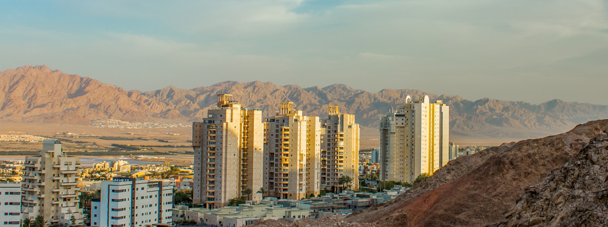 Cityscape in Afghanistan with tall residential buildings set against a backdrop of rugged mountains at sunset, highlighting the contrast between urban development and natural desert terrain.