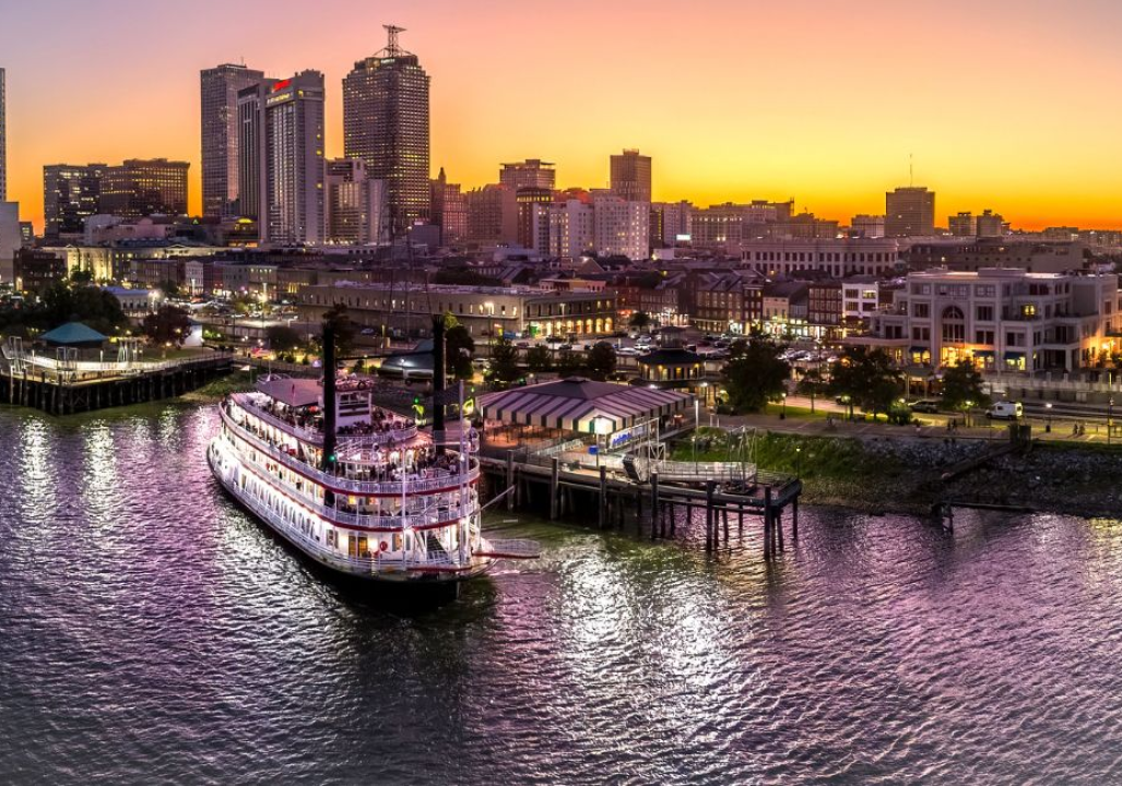 Riverboat docked at a bustling city waterfront during a vibrant sunset. Tall buildings lit up, and the sky is a gradient of golden to purple hues.