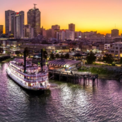 Riverboat docked at a bustling city waterfront during a vibrant sunset. Tall buildings lit up, and the sky is a gradient of golden to purple hues.