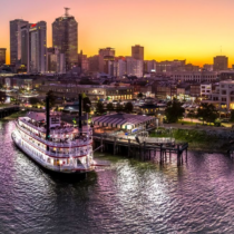 Riverboat docked at a bustling city waterfront during a vibrant sunset. Tall buildings lit up, and the sky is a gradient of golden to purple hues.