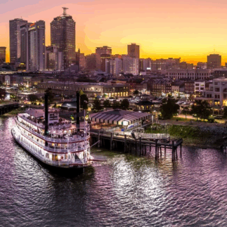 Riverboat docked at a bustling city waterfront during a vibrant sunset. Tall buildings lit up, and the sky is a gradient of golden to purple hues.