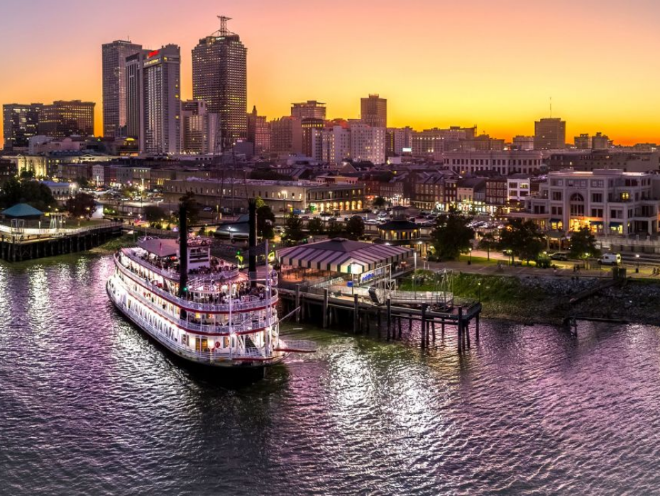 Riverboat docked at a bustling city waterfront during a vibrant sunset. Tall buildings lit up, and the sky is a gradient of golden to purple hues.