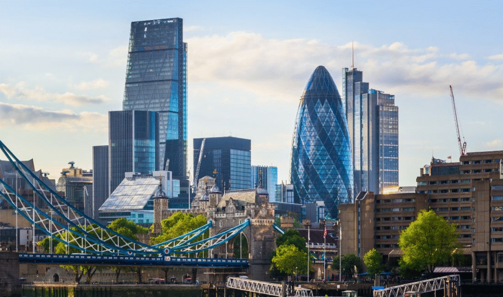 Skyline of London featuring modern skyscrapers like the Gherkin and the Leadenhall Building, with Tower Bridge in the foreground under a blue sky.