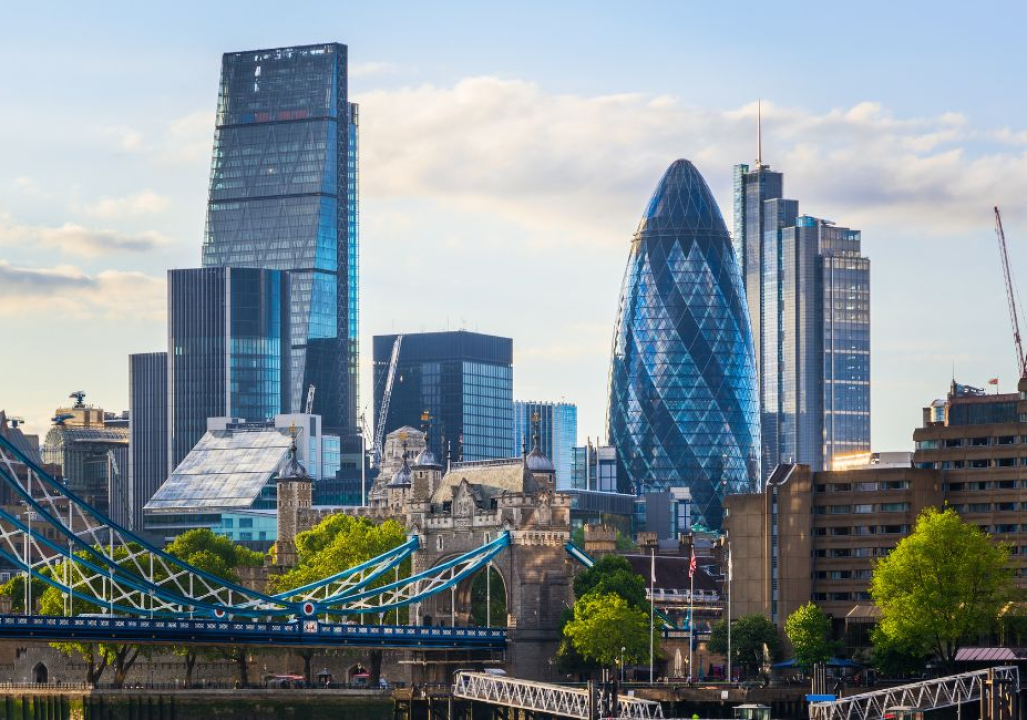 Skyline of London featuring modern skyscrapers like the Gherkin and the Leadenhall Building, with Tower Bridge in the foreground under a blue sky.