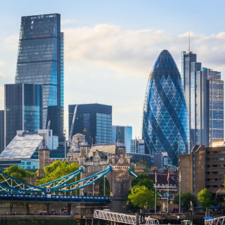 Skyline of London featuring modern skyscrapers like the Gherkin and the Leadenhall Building, with Tower Bridge in the foreground under a blue sky.