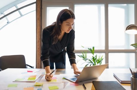 A woman leans over a desk, focusing intently on her laptop. Papers with colorful sticky notes are scattered around. The room is well-lit, conveying a busy work atmosphere.