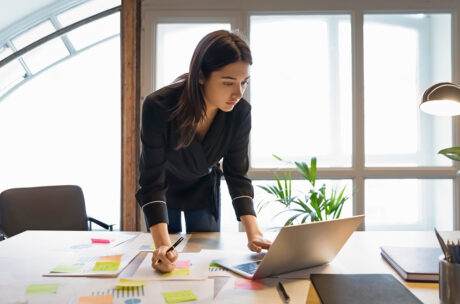 A woman leans over a desk, focusing intently on her laptop. Papers with colorful sticky notes are scattered around. The room is well-lit, conveying a busy work atmosphere.