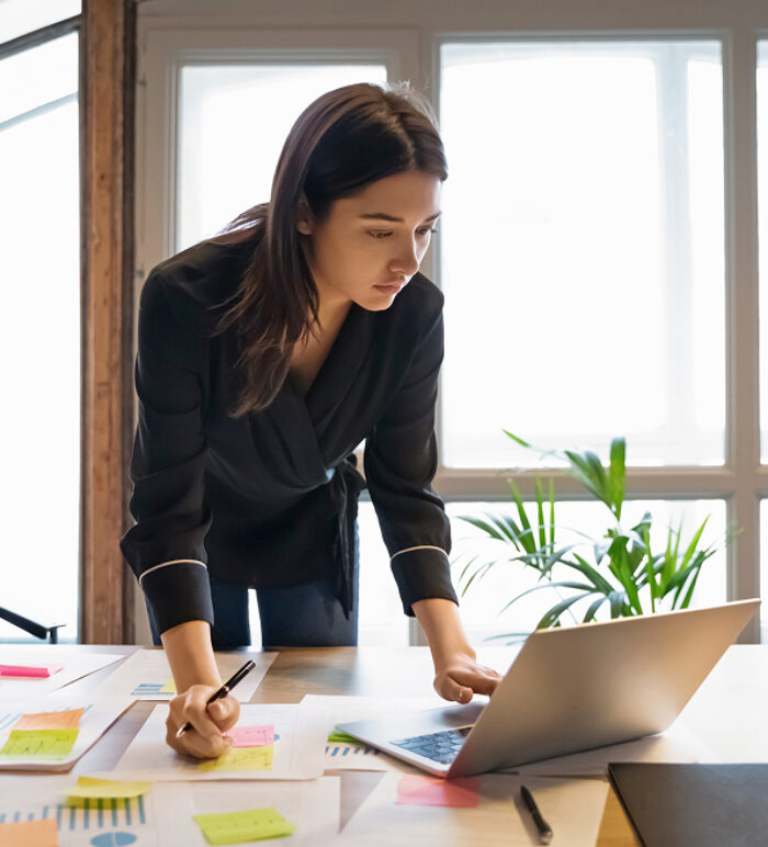A woman leans over a desk, focusing intently on her laptop. Papers with colorful sticky notes are scattered around. The room is well-lit, conveying a busy work atmosphere.