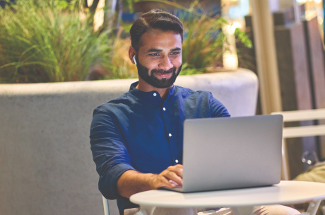Man in a blue shirt, smiling while using a laptop at a white table in a modern, outdoor setting. He wears wireless earbuds, conveying positivity.