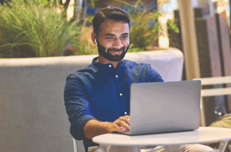 Man in a blue shirt, smiling while using a laptop at a white table in a modern, outdoor setting. He wears wireless earbuds, conveying positivity.
