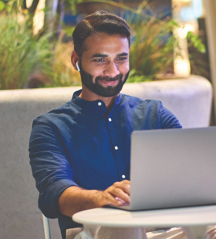 Man in a blue shirt, smiling while using a laptop at a white table in a modern, outdoor setting. He wears wireless earbuds, conveying positivity.