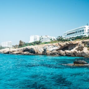 coastal scene in Cyprus, featuring clear turquoise waters, rocky cliffs, and white modern buildings